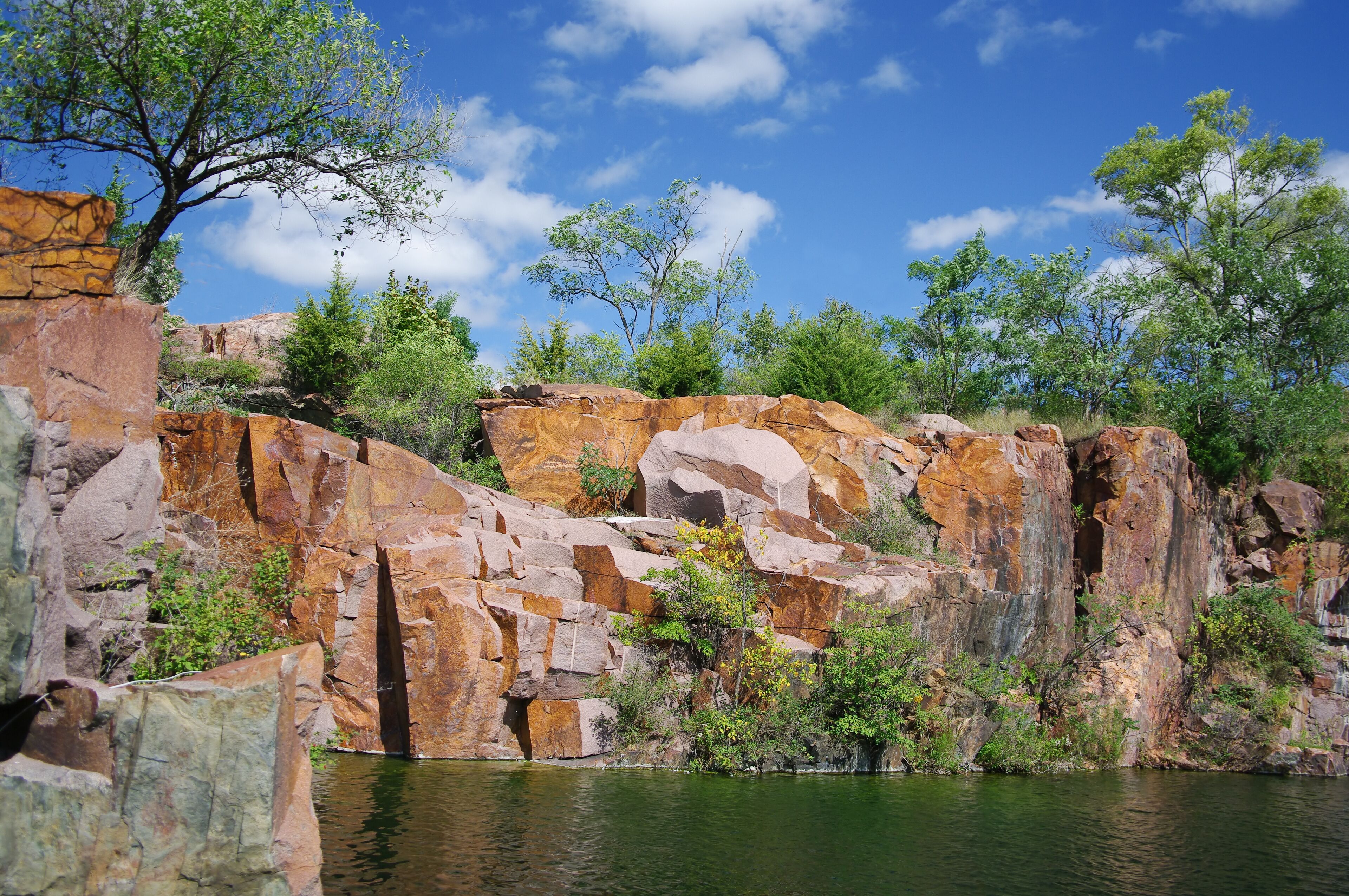 A rocky cliff with a river at its base, surrounded by greenery and trees.