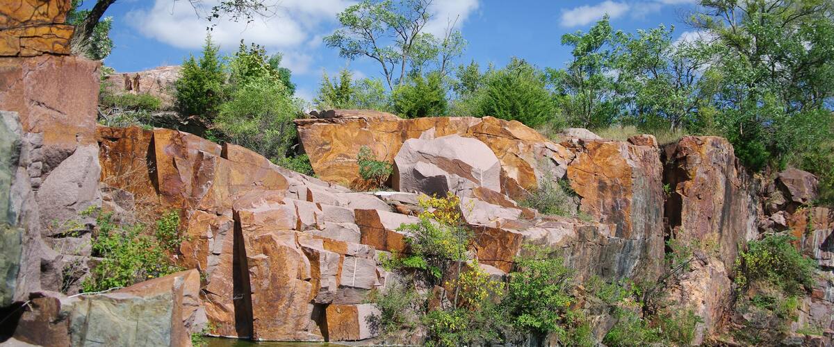 A rocky cliff with a river at its base, surrounded by greenery and trees.