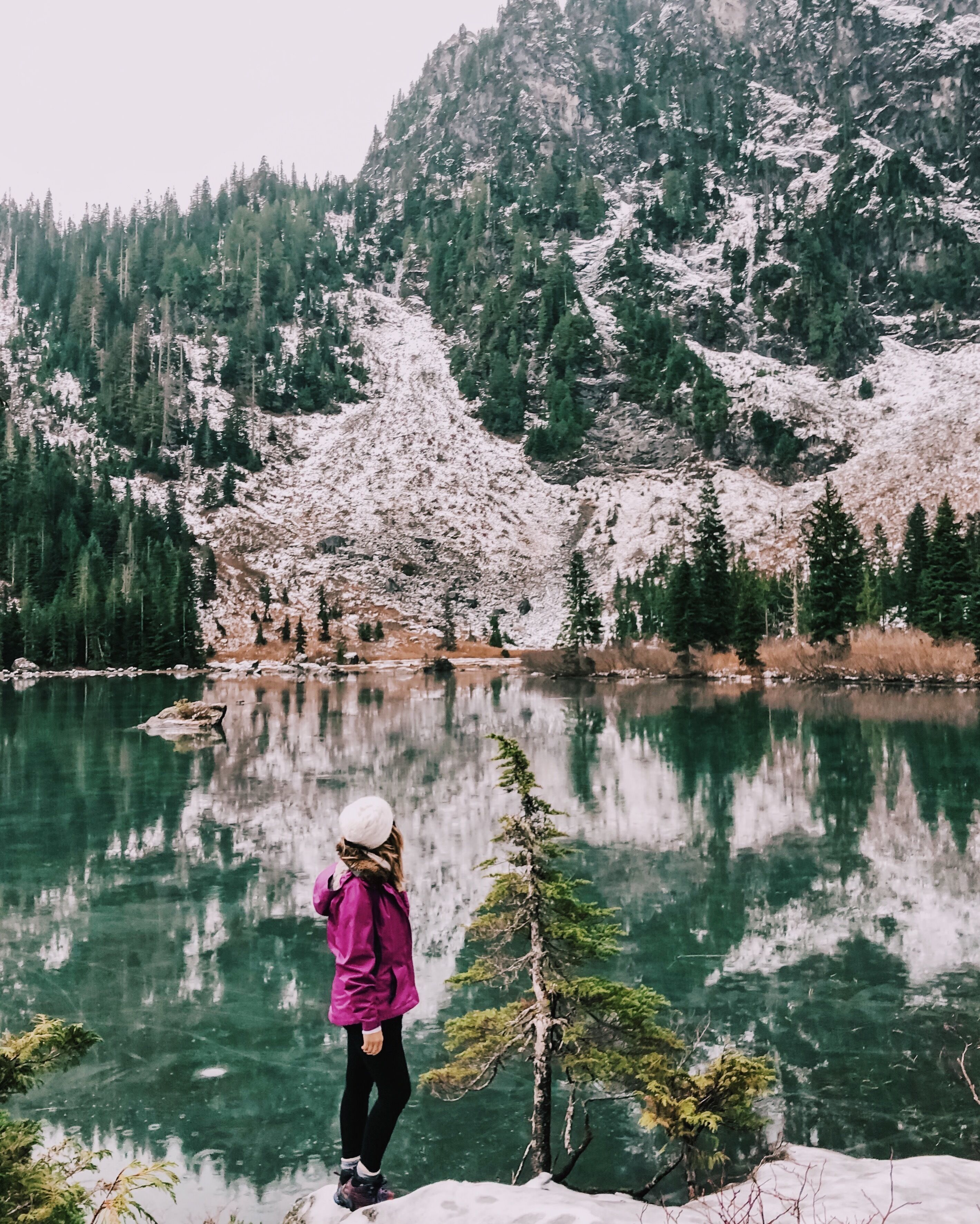 A person in a purple jacket and white hat standing by a clear lake with snow-covered mountains in the background.