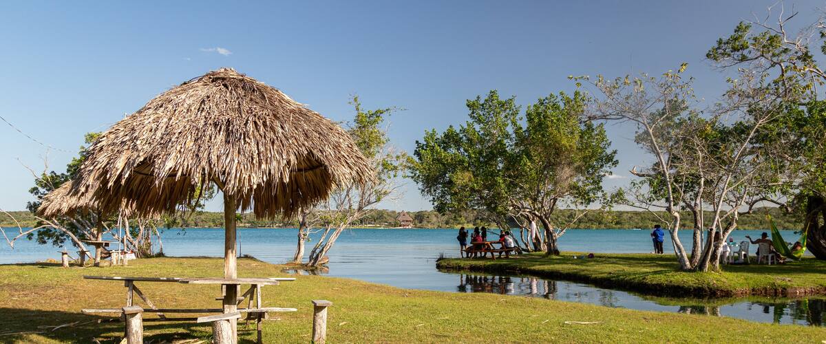 A grassy area with a thatched-roof pavilion, a calm lake, and people relaxing by the water.
