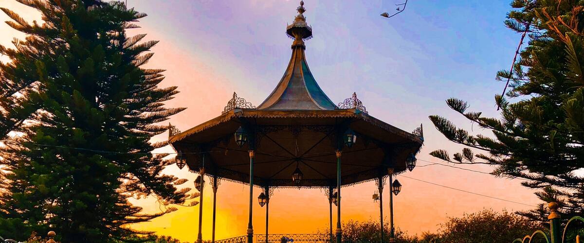 A gazebo with a pointed roof and ornate railings, surrounded by a paved walkway and trees.