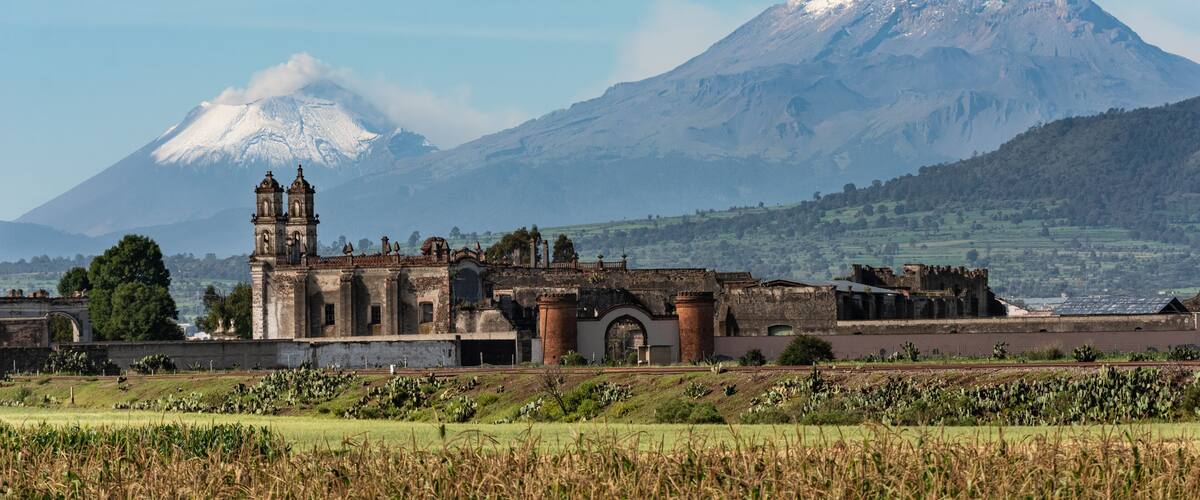 A historic church with a bell tower, a snow-capped mountain, and a green valley.