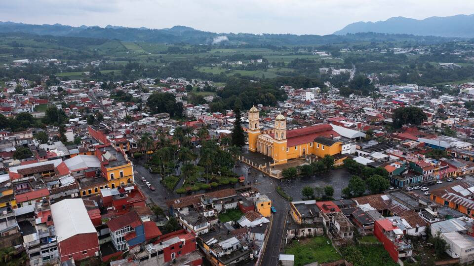 A cityscape with a prominent yellow church with two towers, surrounded by densely packed buildings and green hills in the background.