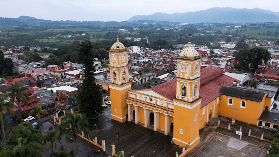 A yellow church with two towers and a dome, surrounded by a cityscape and mountains in the background.