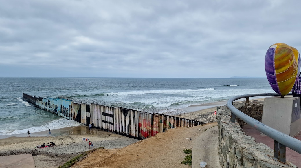 A coastal scene with a graffiti-covered pier extending into the sea, a sandy beach, and a colorful sculpture.