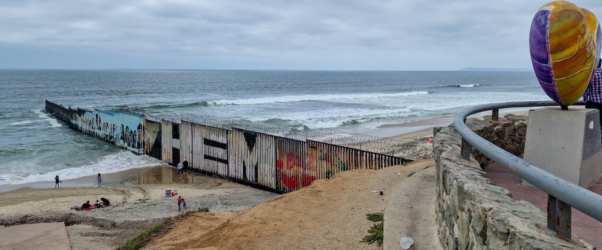 A coastal scene with a graffiti-covered pier extending into the sea, a sandy beach, and a colorful sculpture.