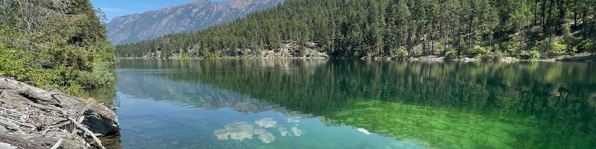 A clear lake with a blue kayak on the shore, surrounded by mountains and dense forest.