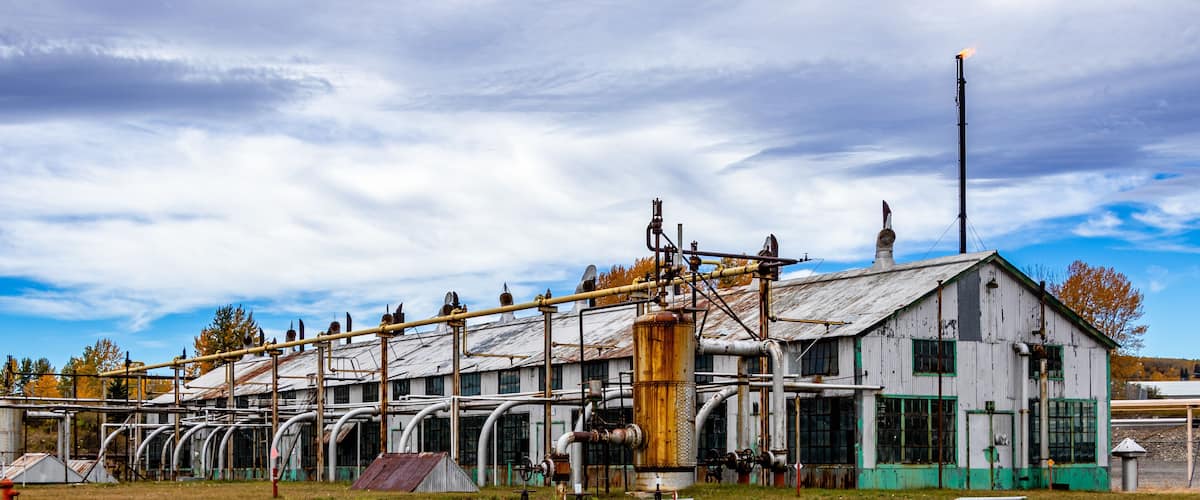 An abandoned industrial complex with rusted machinery, a large cylindrical tank, and a weathered building with green trim.