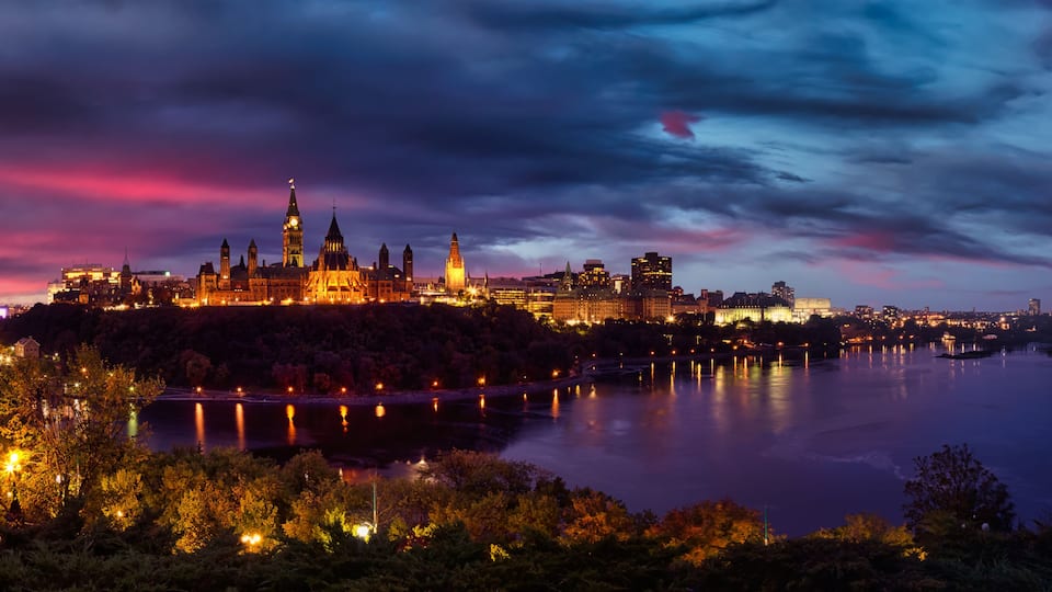 A cityscape at dusk with illuminated buildings, a prominent clock tower, and a calm river reflecting the lights.