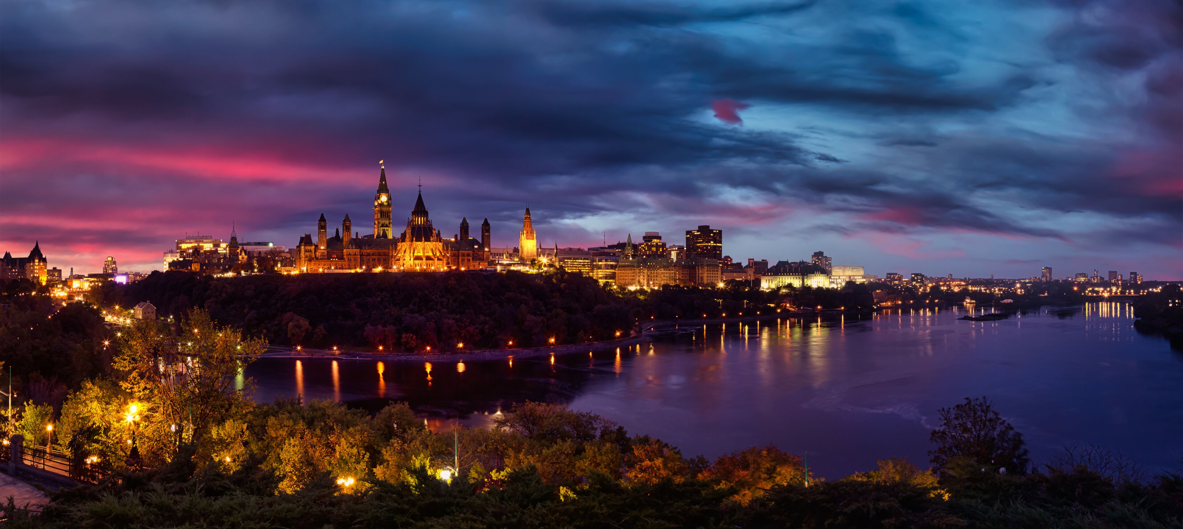 A cityscape at dusk with illuminated buildings, a prominent clock tower, and a calm river reflecting the lights.