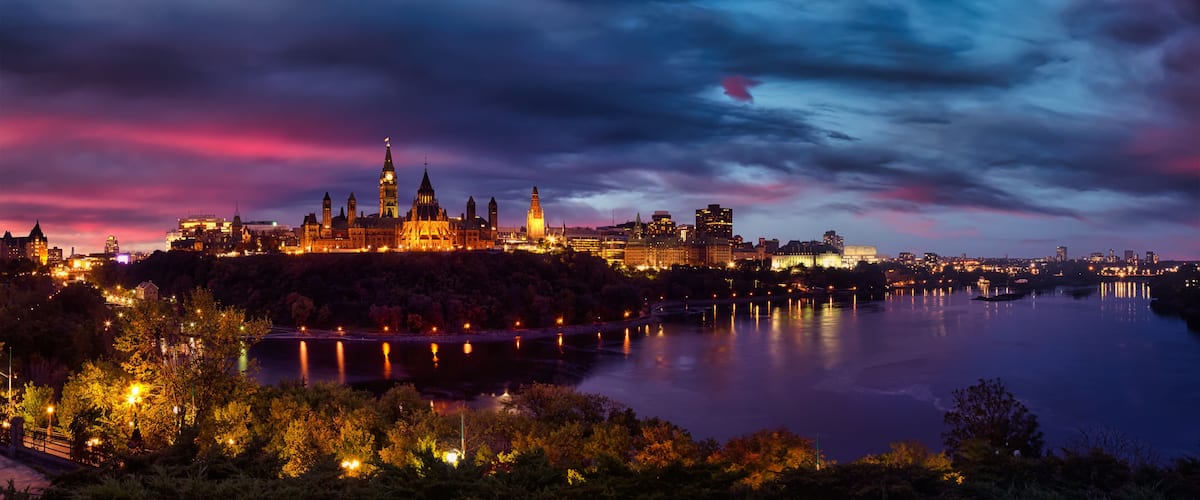 A cityscape at dusk with illuminated buildings, a prominent clock tower, and a calm river reflecting the lights.