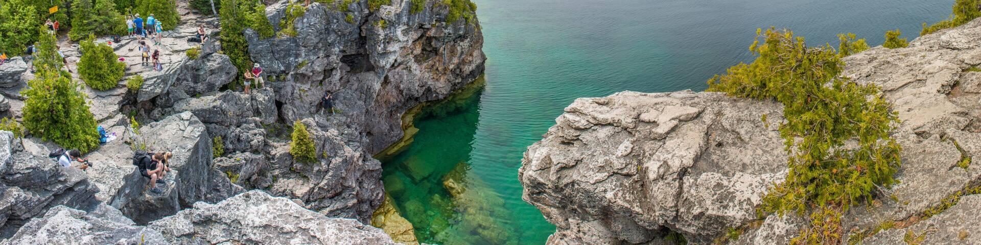 A rocky coastline with clear water, dense forest, and a group of people on the cliff.