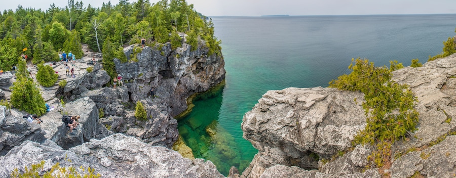 A rocky coastline with clear water, dense forest, and a group of people on the cliff.