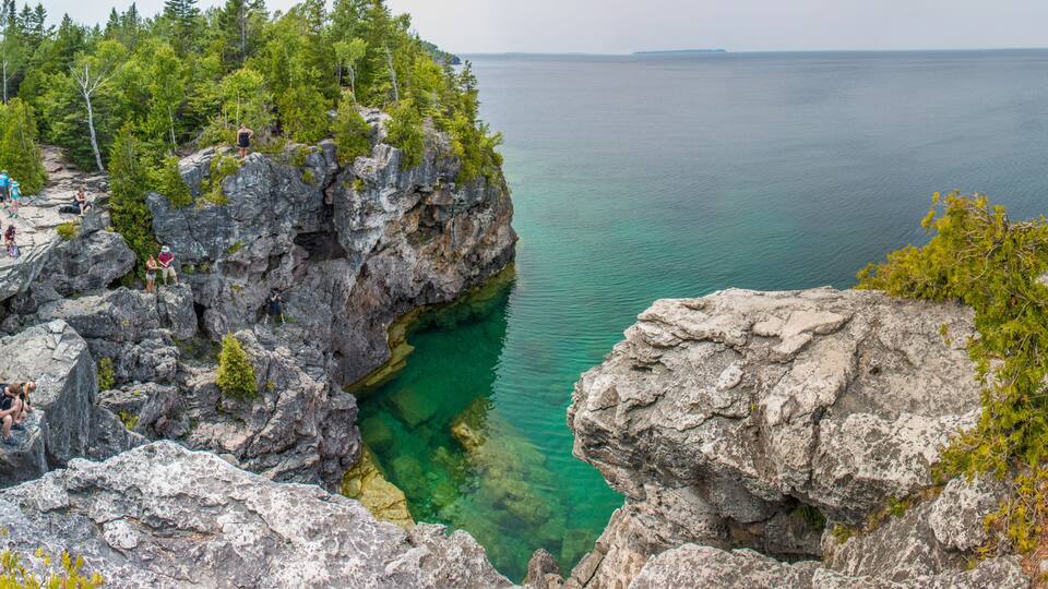 A rocky coastline with clear water, dense forest, and a group of people on the cliff.
