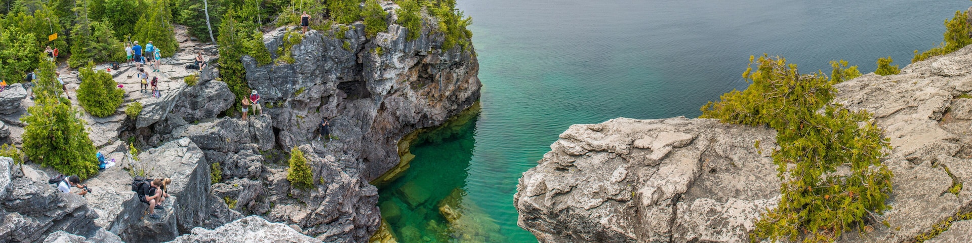 A rocky coastline with clear water, dense forest, and a group of people on the cliff.