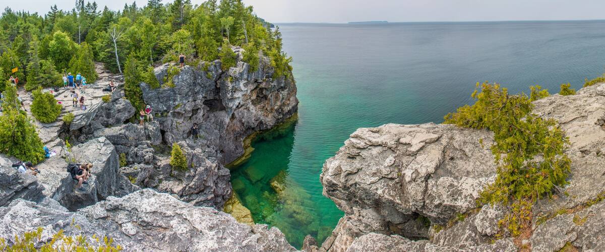 A rocky coastline with clear water, dense forest, and a group of people on the cliff.