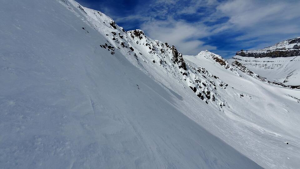 A snow-covered mountain slope with rocky outcrops and a clear blue sky.