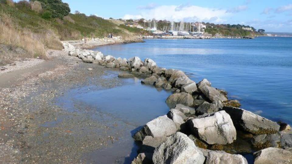 A rocky shoreline with a calm sea, a distant sailboat, and a hilly landscape with buildings.