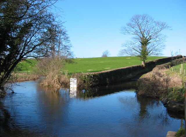 Impassable ford of the River Weaver. Woodcotthill Road apparently once forded the River Weaver (left to right foreground) at this point; however, as the depth gauge (4 ft) reveals, the ford is impassable to vehicles. The road is still marked as a ford to the north but as a no-through road to the south. A footbridge (out of view on right; see this image) allows people and bicycles to pass with dry feet. The River Weaver is here the boundary between the Cheshire civil parishes of Newhall and Wrenbury cum Frith.