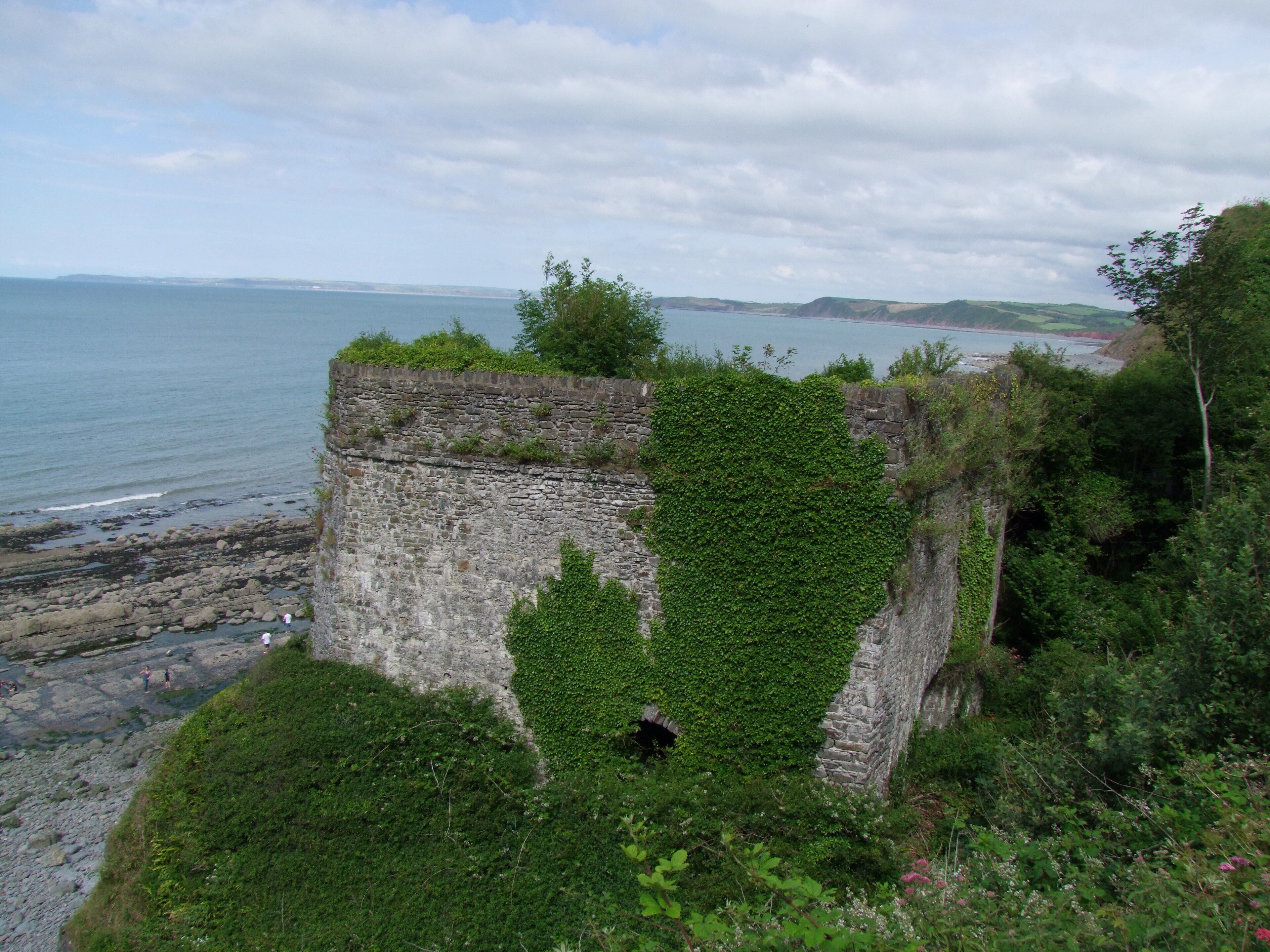 A coastal stone structure with greenery, overlooking the sea and distant hills.
