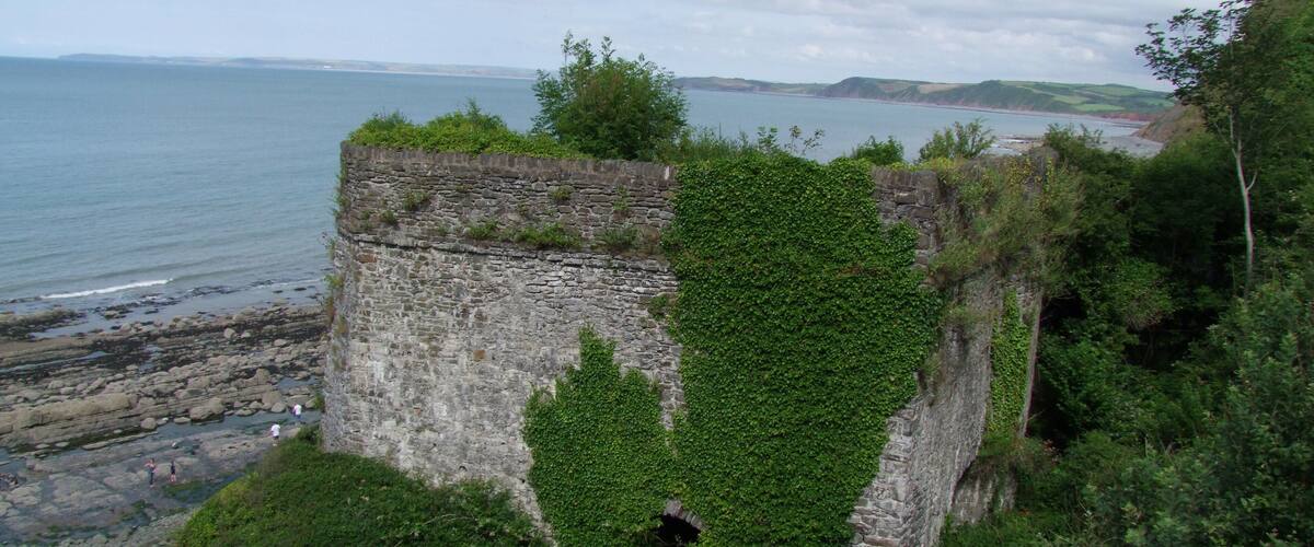 A coastal stone structure with greenery, overlooking the sea and distant hills.