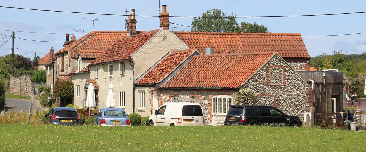 A rural house with a red-tiled roof, surrounded by a grassy field and parked cars.