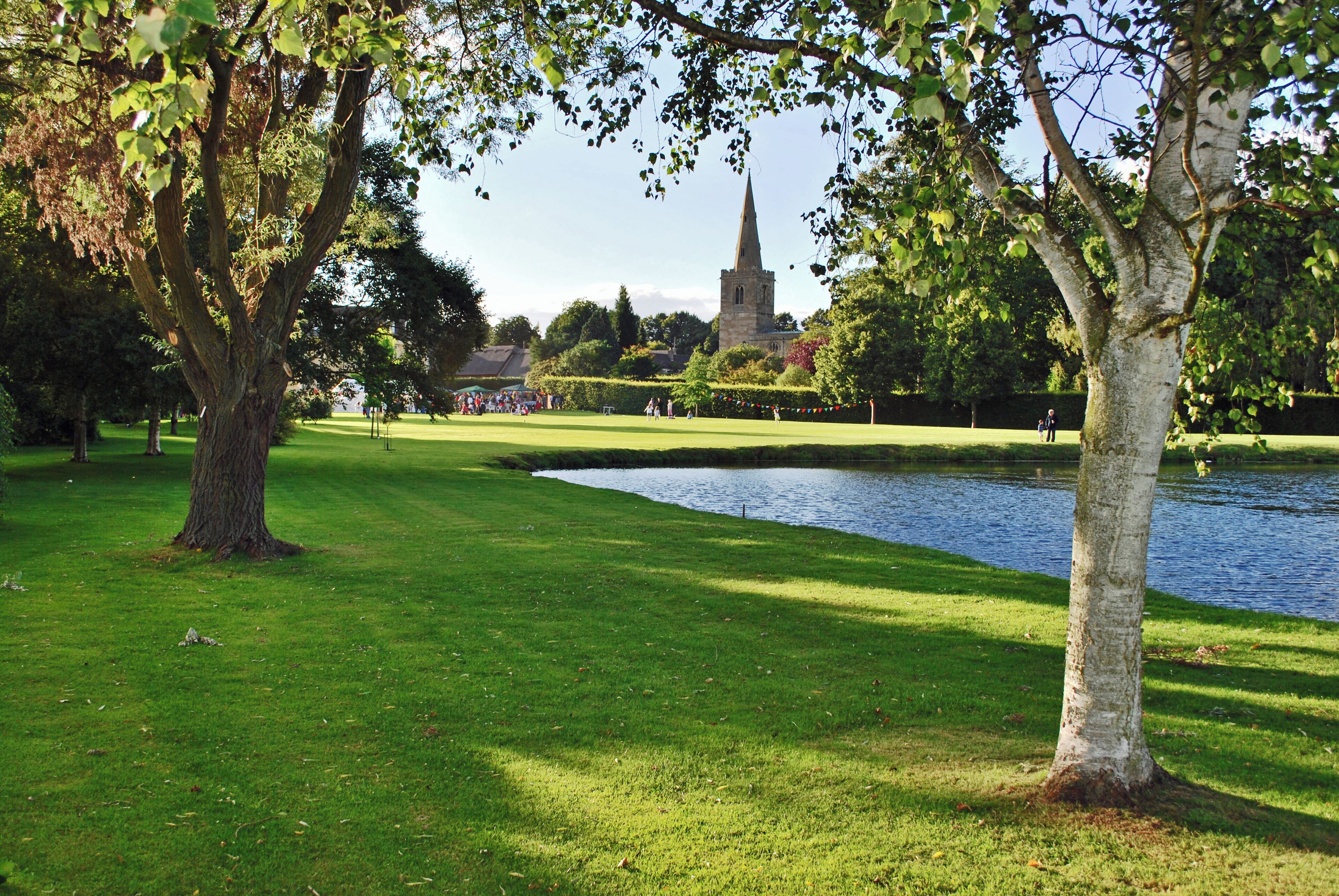 A park with a pond, trees, and a church steeple in the background.