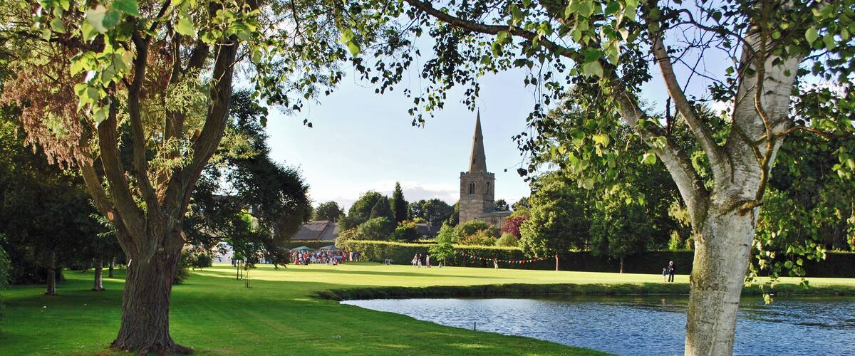 A park with a pond, trees, and a church steeple in the background.