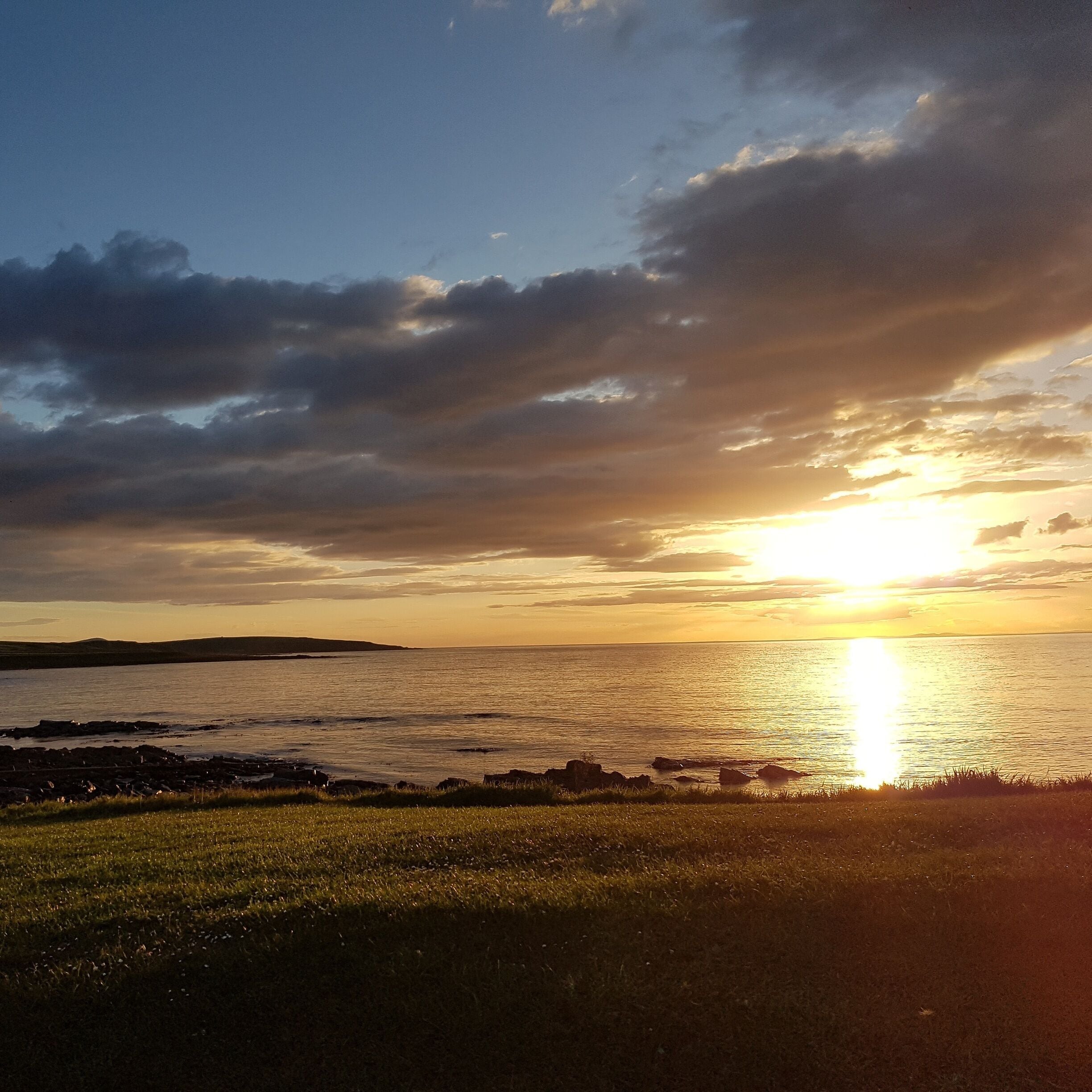 A sunset over a calm sea with a grassy foreground and rocky shoreline.