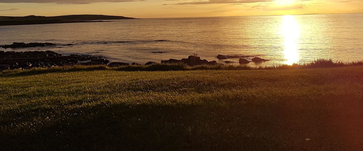 A sunset over a calm sea with a grassy foreground and rocky shoreline.