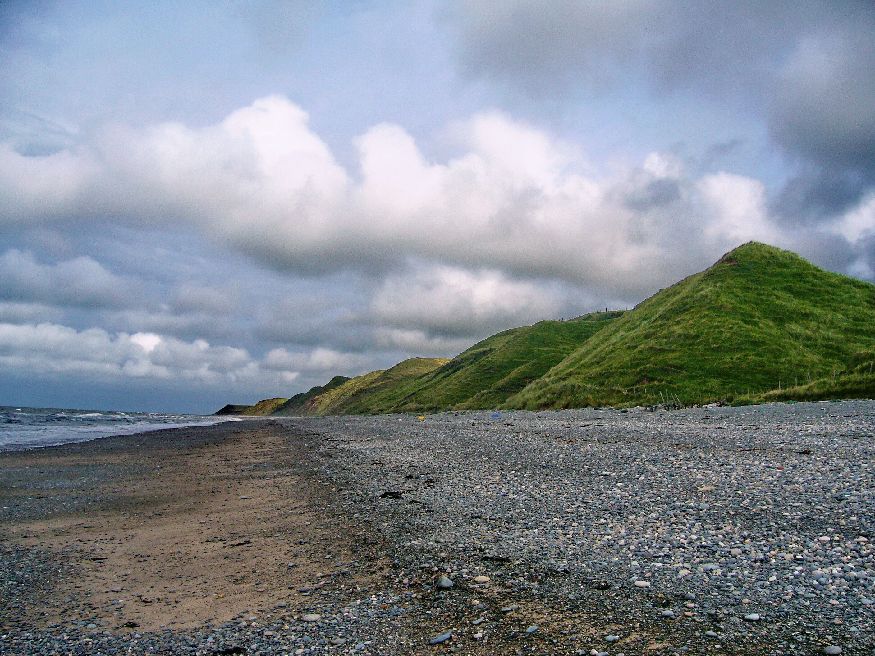 A pebbly beach with a grassy hill in the background under a cloudy sky.