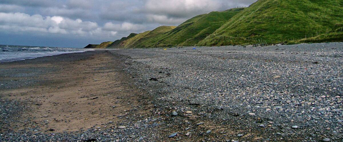 A pebbly beach with a grassy hill in the background under a cloudy sky.