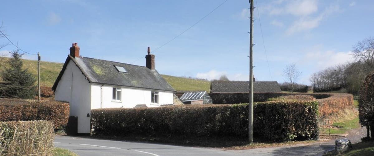 A rural road with a white house on the left, surrounded by hedges and a grassy hill in the background.