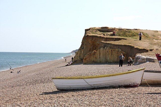 A pebbled beach with a boat, people, and a cliff.