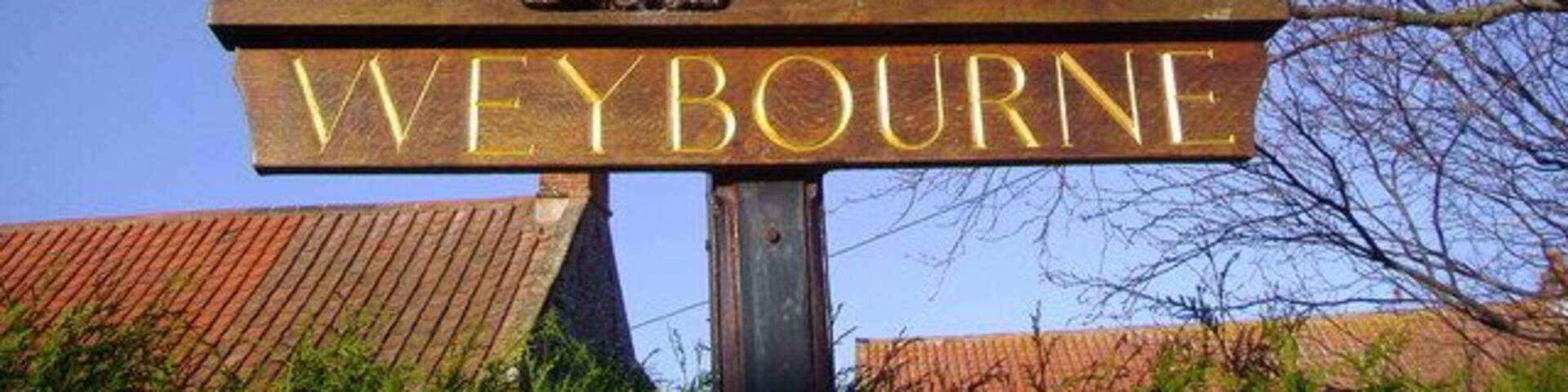 A wooden sign with 'WEYBOURNE' and a horse-drawn carriage scene.