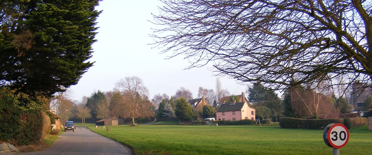 A rural road with a 30 mph speed limit sign, surrounded by trees and houses.