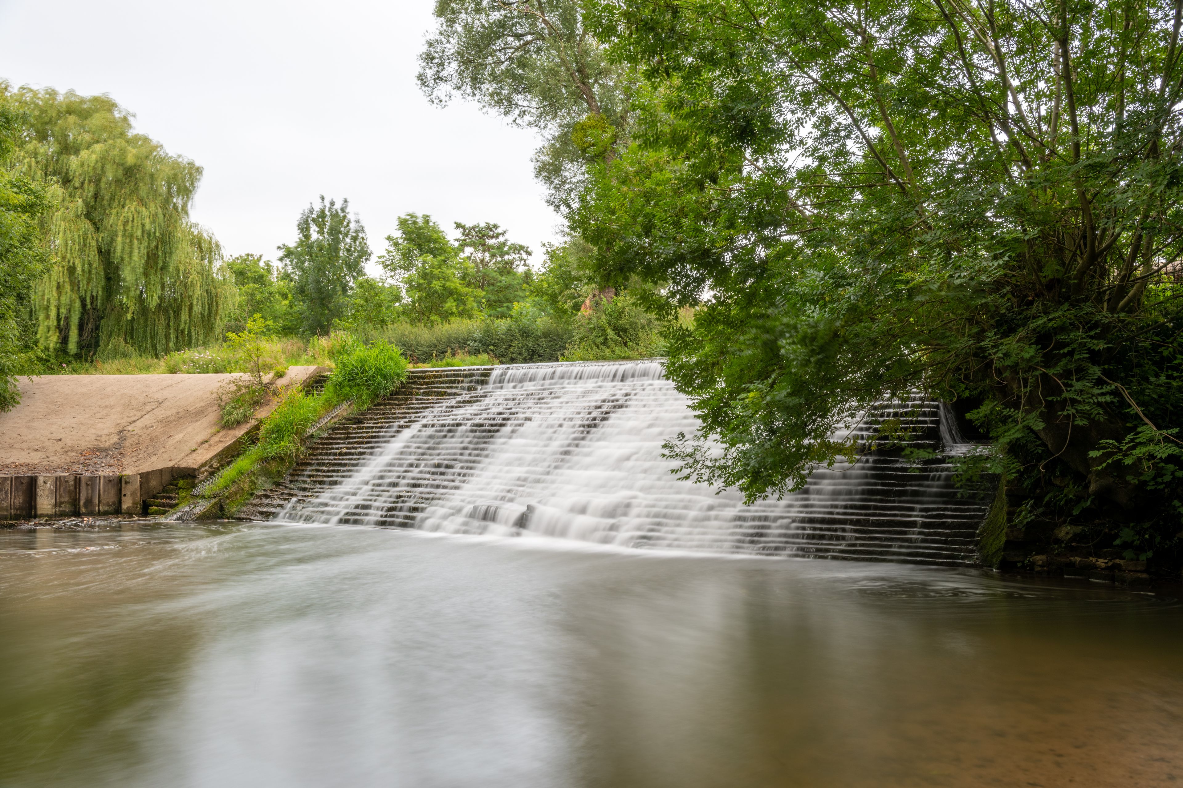 A waterfall cascading over a concrete spillway into a river, surrounded by lush greenery and trees.