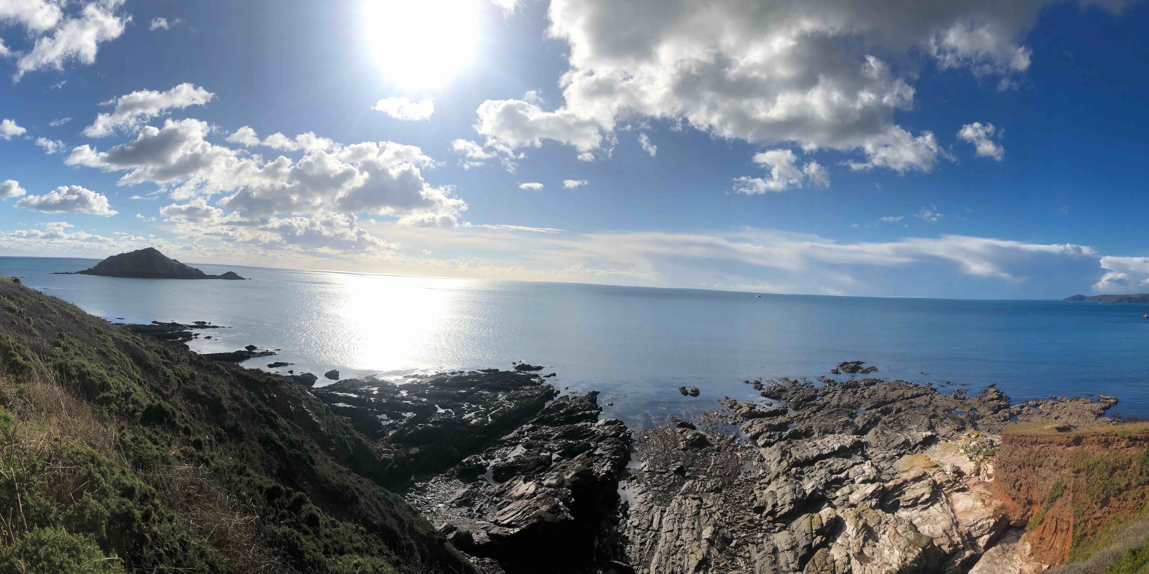 A coastal landscape with rocky cliffs, a calm sea, and a distant island under a partly cloudy sky.