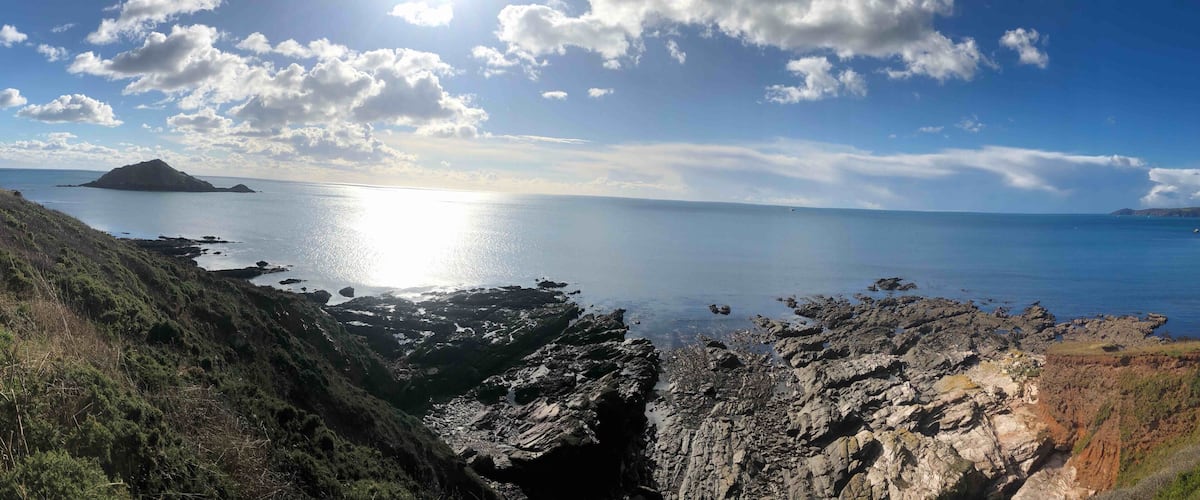 A coastal landscape with rocky cliffs, a calm sea, and a distant island under a partly cloudy sky.