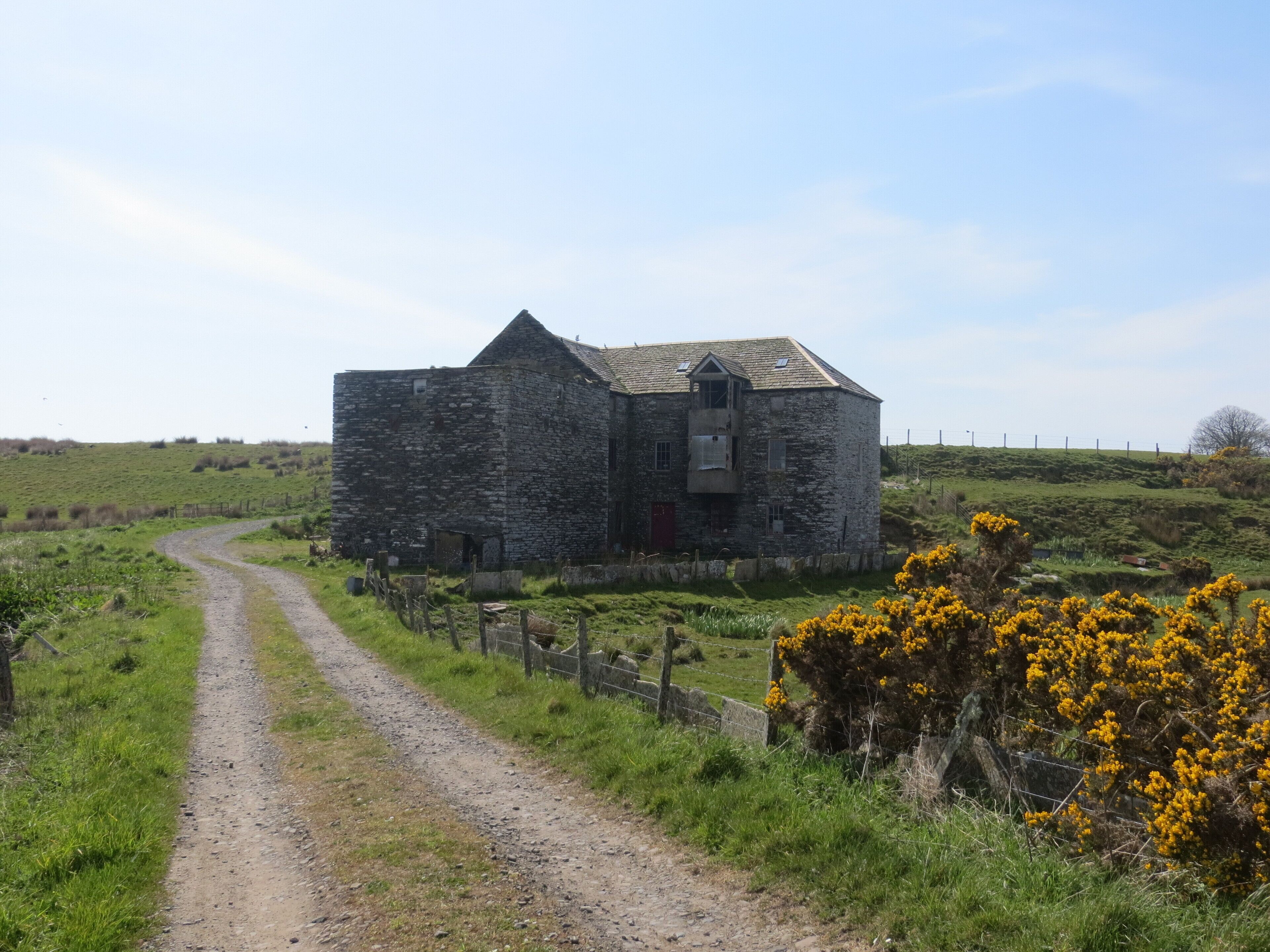 A stone building with a red door, surrounded by a fenced field and a dirt path.