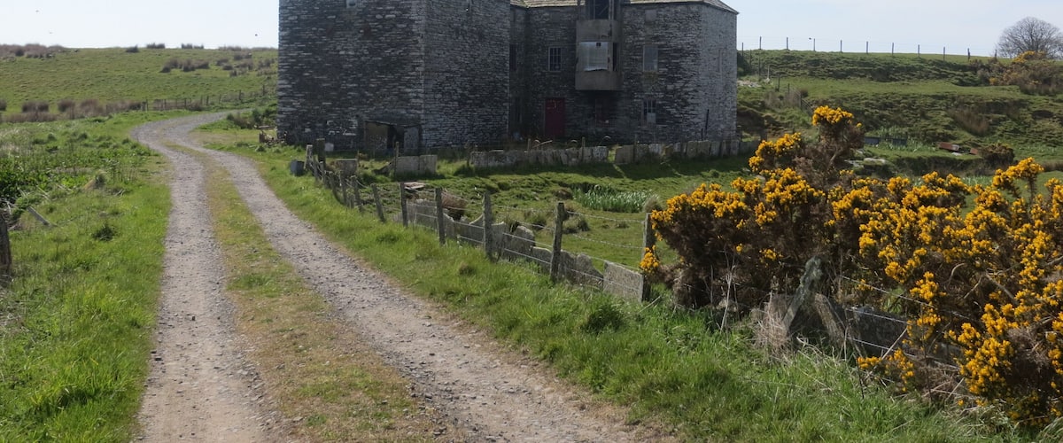 A stone building with a red door, surrounded by a fenced field and a dirt path.