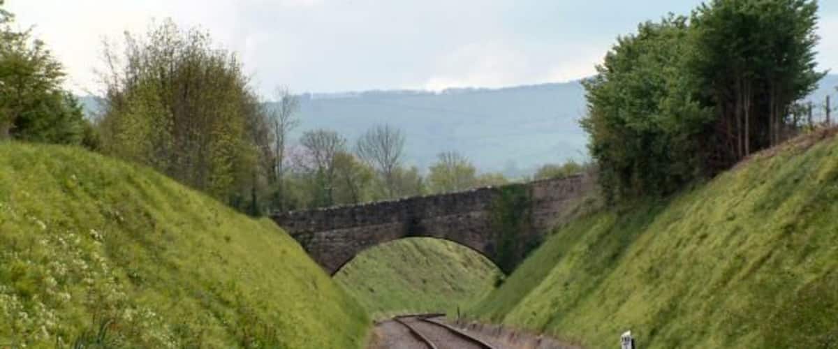 Railway tracks curving through a green landscape with a stone bridge in the distance.
