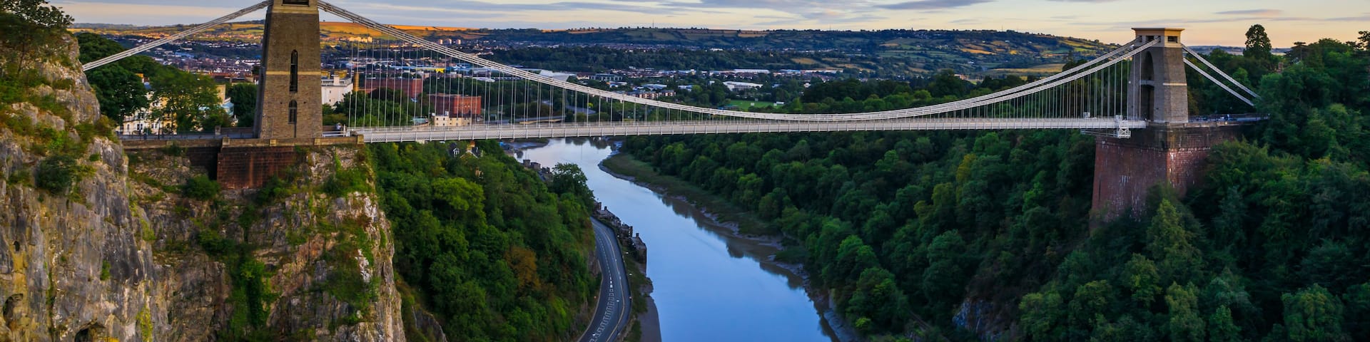 A suspension bridge spanning a river, surrounded by lush greenery and a clear sky.