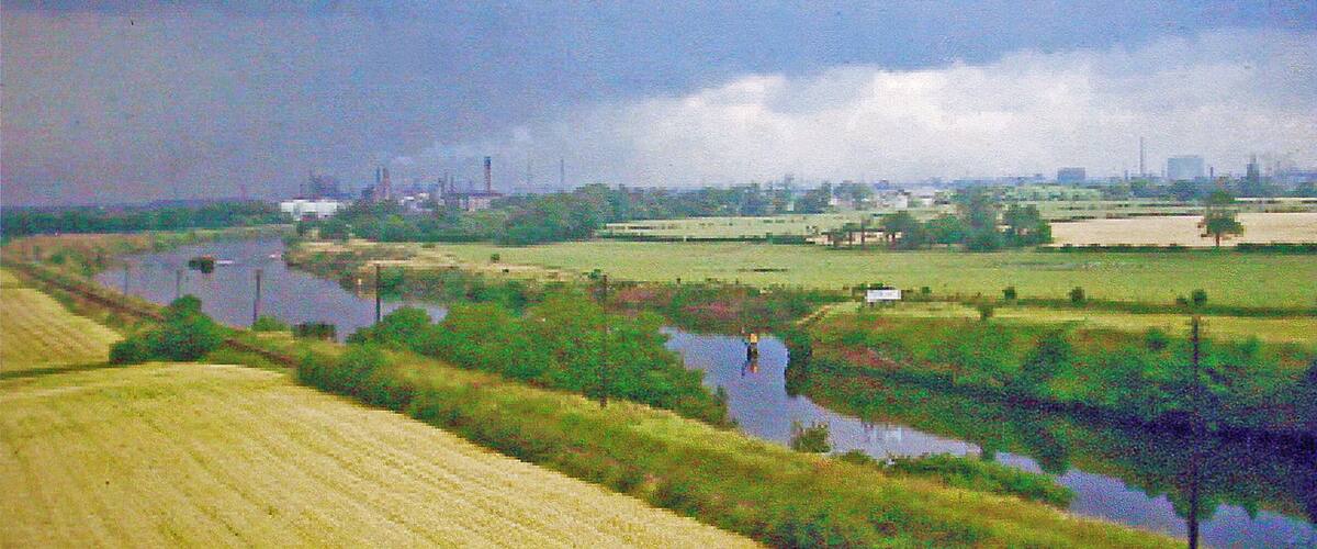 A river meandering through a rural landscape with fields, a small body of water, and distant industrial structures.