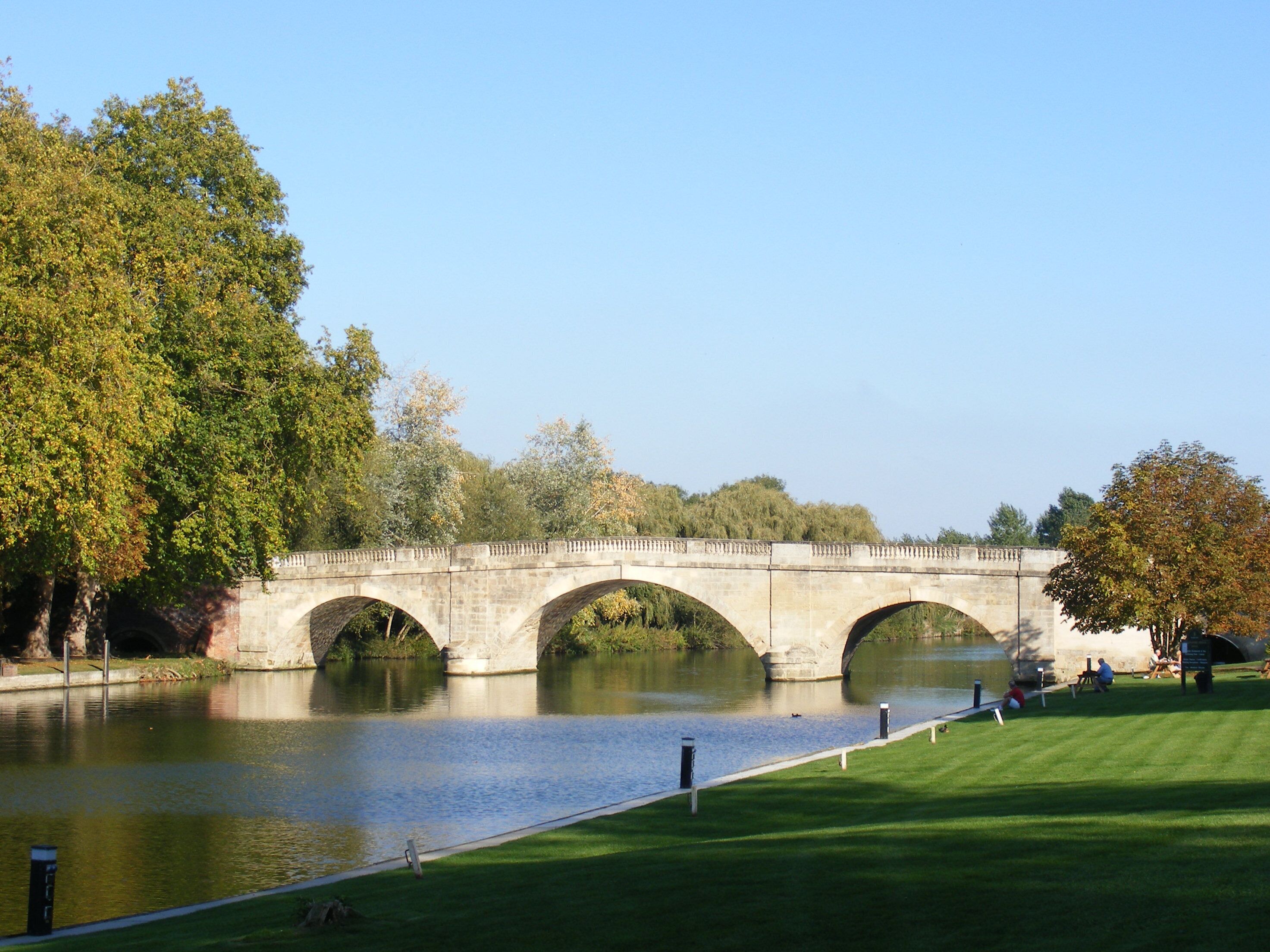A stone bridge over a calm river, surrounded by lush trees and a well-maintained grassy area.