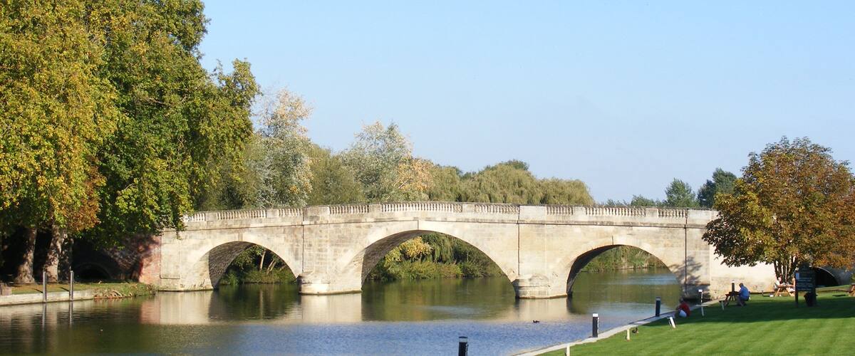 A stone bridge over a calm river, surrounded by lush trees and a well-maintained grassy area.