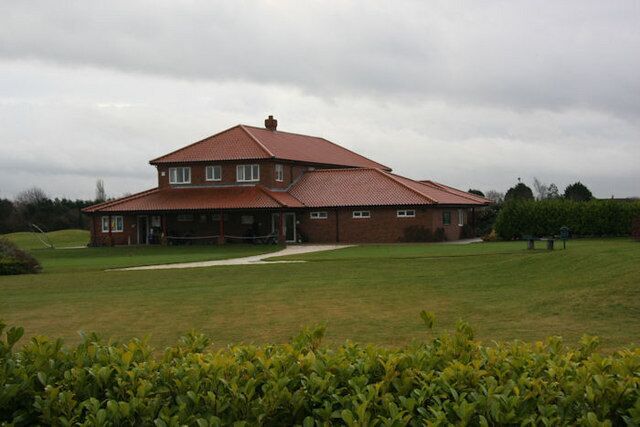 A large brick house with a red roof, surrounded by a grassy field and a hedge.