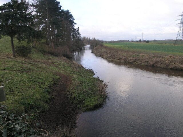 A narrow waterway flanked by grassy banks and trees, with distant power lines and a cloudy sky.