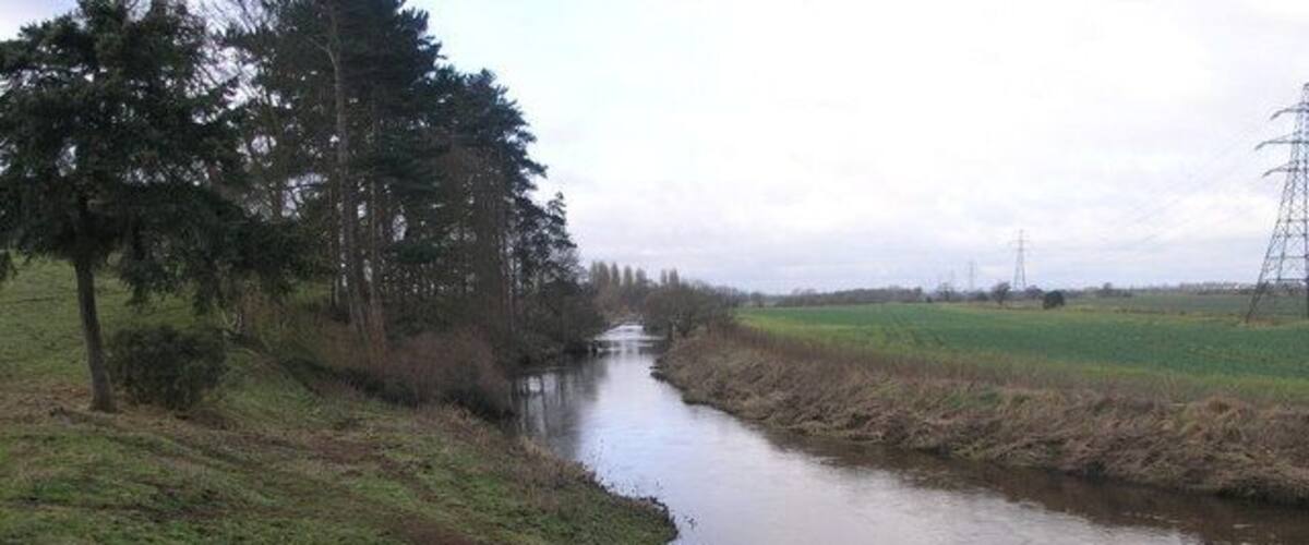 A narrow waterway flanked by grassy banks and trees, with distant power lines and a cloudy sky.