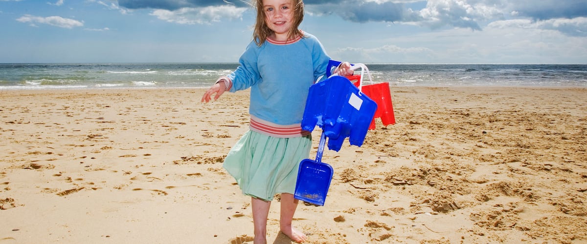 A child on a sandy beach with a blue and red shovel, holding a blue bucket, and a red beach chair in the background.